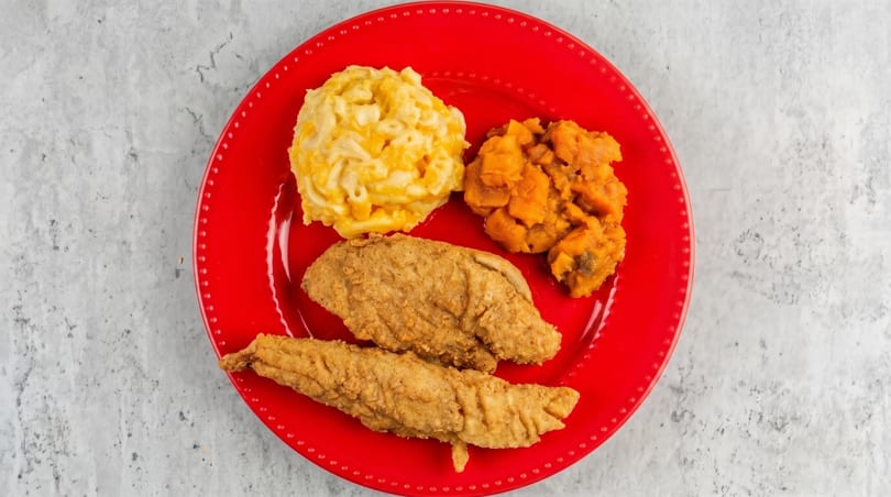 Fried fish fillets, macaroni and cheese, and candied yams on a bright red plate.