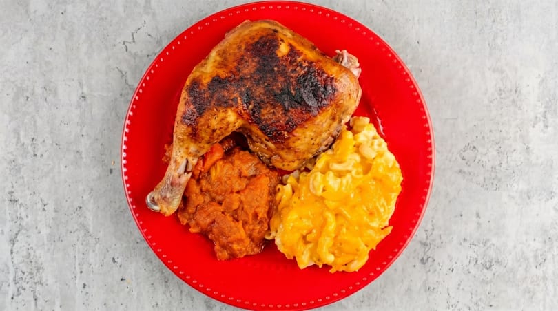Grilled chicken leg and thigh with mashed sweet potatoes and macaroni and cheese on a red plate.