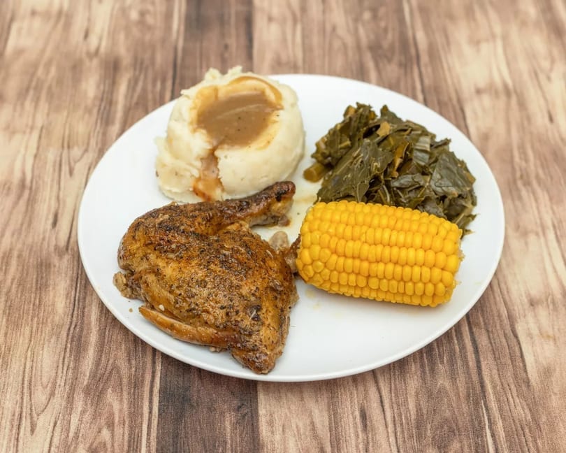 A plate of roasted chicken, mashed potatoes with gravy, collard greens, and corn on the cob.